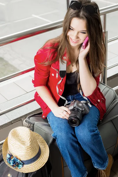 Jovem mulher atraente em casaco vermelho sentado em malas no terminal ou estação de trem e falando ao telefone . — Fotografia de Stock