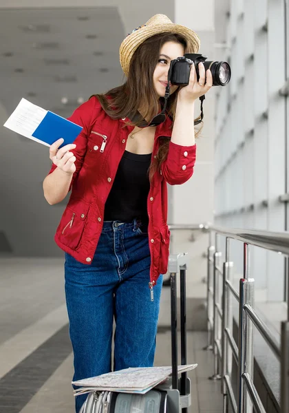 Joven atractiva mujer en un abrigo rojo y sombrero de pie en la terminal o en la estación con una maleta y fotografías — Foto de Stock