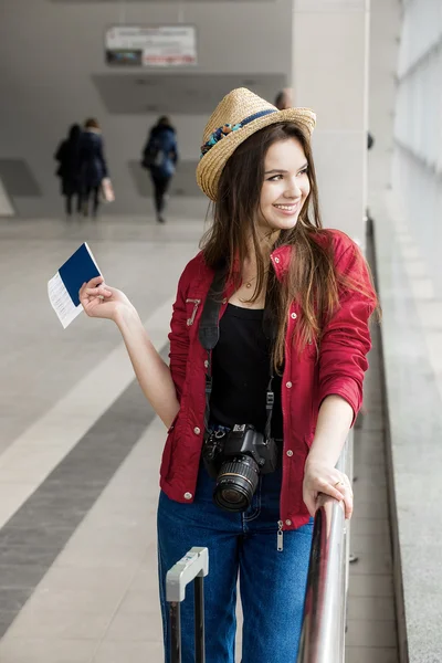 Mujer joven con un abrigo rojo y sombrero de pie en la terminal o en la estación con una maleta y pasaporte — Foto de Stock