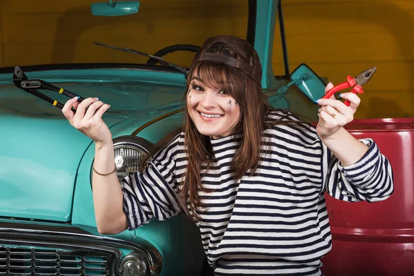cheerful woman sits in the garage near the retro car with tools. Girl holding pliers and an adjustable wrench