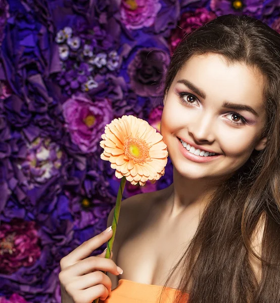 Mulher morena com uma maquiagem suave que olha para a câmera enquanto segura a flor perto do rosto em um fundo floral . — Fotografia de Stock