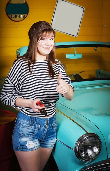 Mulher morena em um boné e uma camisola listrada mostrando polegar para cima e segurando ferramentas na garagem perto do carro retro — Fotografia de Stock