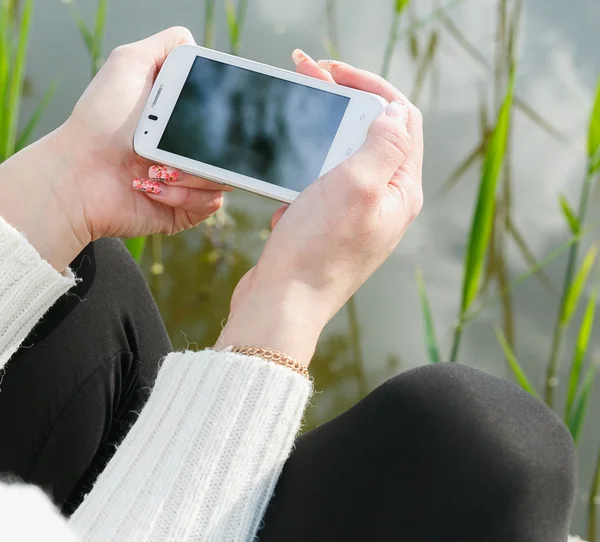 Piękna młoda kobieta za pomocą telefonu komórkowego, siedząc na trawie. Lato zdjęcie. Przybrzeżnych widok. — Zdjęcie stockowe