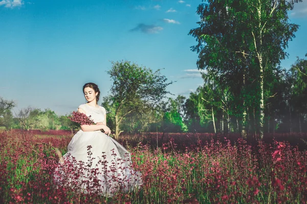 Bela noiva em um campo de flores. A menina em um vestido branco com um buquê em um campo de verão ao pôr do sol — Fotografia de Stock