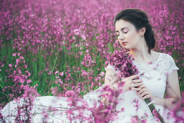 Belle mariée dans un champ de fleurs. La fille dans une robe blanche avec un bouquet dans un champ d'été au coucher du soleil — Photo