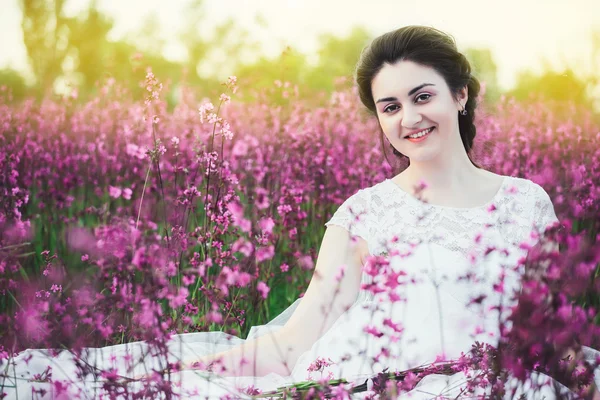 Belle mariée dans un champ de fleurs. La fille dans une robe blanche avec un bouquet dans un champ d'été au coucher du soleil — Photo