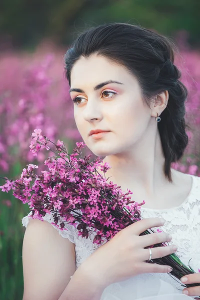 Hermosa novia en un campo de flores. La muchacha en el vestido blanco con el ramo en el campo veraniego al atardecer —  Fotos de Stock