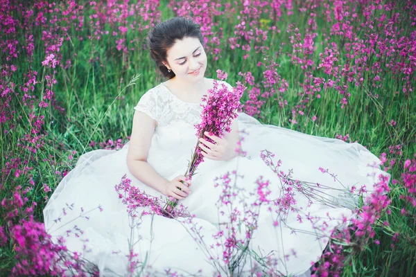Hermosa novia en un campo de flores. La muchacha en el vestido blanco con el ramo en el campo veraniego al atardecer —  Fotos de Stock