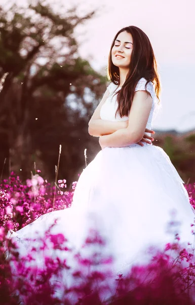 Hermosa novia en un campo de flores. La chica en un vestido blanco con un ramo en un campo de verano al atardecer. Mujer joven en vestido de novia al aire libre . — Foto de Stock