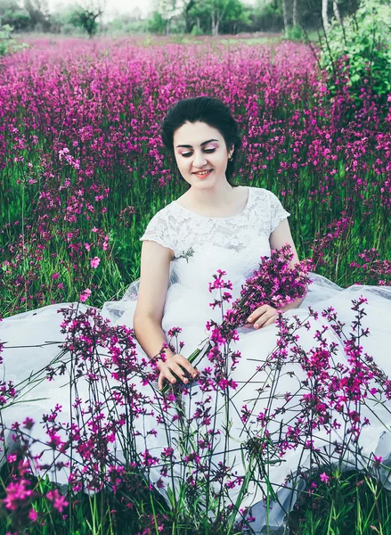 Όμορφη νύφη σε ένα πεδίο λουλούδι. Το κορίτσι σε ένα λευκό φόρεμα με μια ανθοδέσμη σε ένα πεδίο καλοκαίρι στο ηλιοβασίλεμα — Φωτογραφία Αρχείου