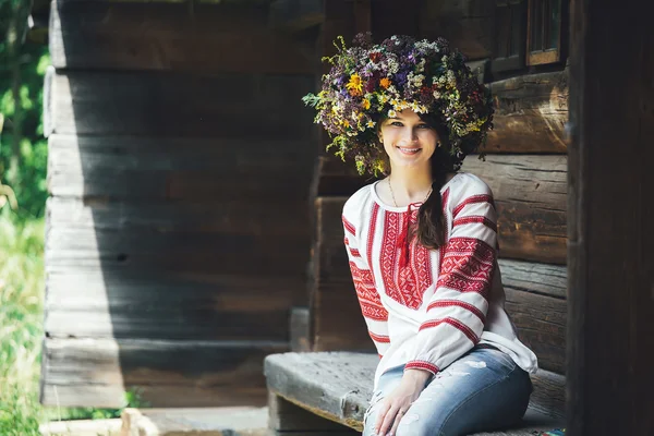 Bela jovem mulher em traje tradicional ucraniano e uma grinalda floral grande senta-se em um banco perto de casa de madeira — Fotografia de Stock
