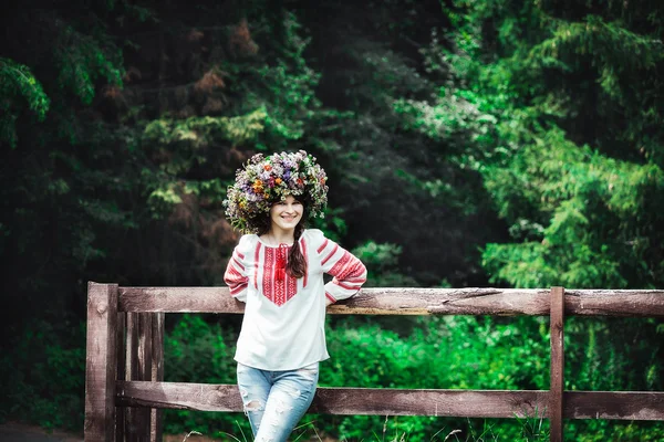 Belle jeune femme en costume traditionnel ukrainien et une grande couronne florale se tient près d'une clôture en bois dans une forêt — Photo