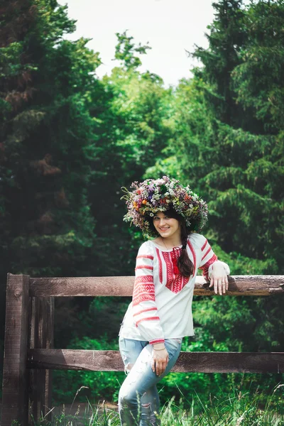 Krásná mladá žena v kroji ukrajinských a velký květinový věnec stojí v blízkosti dřevěného plotu v lese — Stock fotografie