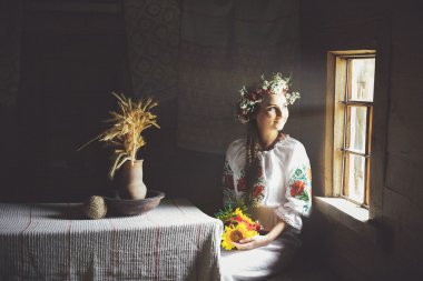 geleneksel elbiseli bir genç Ukraynalı kadın karanlık bir odada ve pencereden dışarı bakarak bir masada oturur.