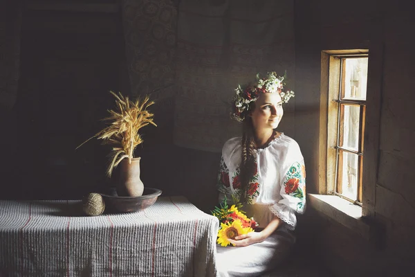 身穿传统服装的年轻乌克兰女子坐在一张桌子在漆黑的房间，看着窗外. — 图库照片