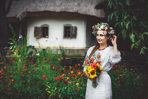 Uma jovem mulher ucraniana em vestido tradicional em pé no quintal em um fundo de uma antiga casa de campo — Fotografia de Stock