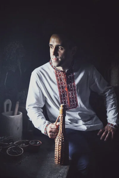 A young Ukrainian man in traditional dress sitting with a bottle in a dark room — Stock Photo, Image