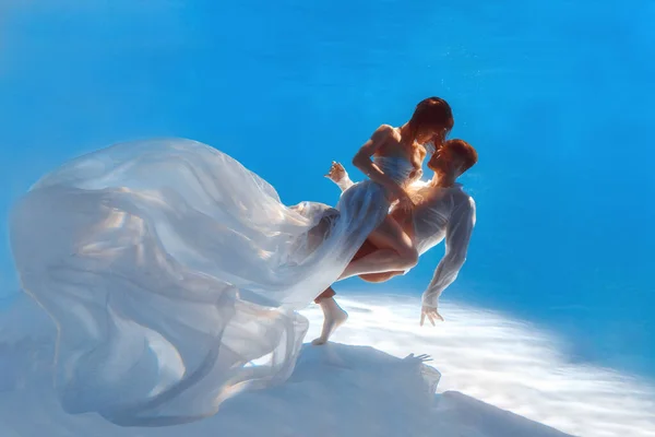 The couple is dancing or hugging in the pool underwater. A girl in a dress with a long train and a guy swim underwater.