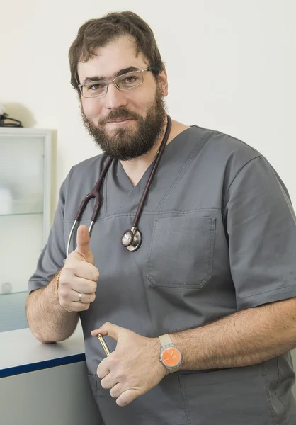 Médico barbudo usando óculos e um manto cinza mostrando polegar — Fotografia de Stock