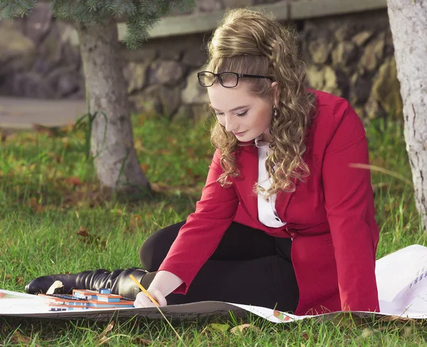 Young beautiful girl in a red jacket sitting on the grass on a large sheet of paper and draw a plan to build a building considering the layout — Zdjęcie stockowe