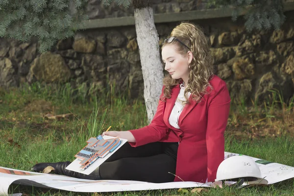 Молодая красивая девушка в красной куртке сидит на траве на большом листе бумаги и нарисовать план строительства здания с учетом макета — стоковое фото