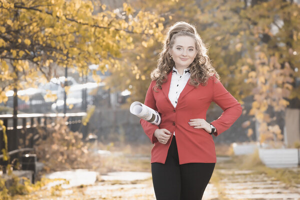 Young woman architect in red jacket and white helmet comes in the autumn on the street and keeps convolution with the drawings. backlight