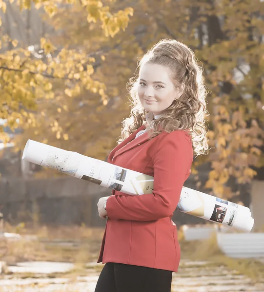 Young woman architect in red jacket and white helmet comes in the autumn on the street and keeps convolution with the drawings. backlight