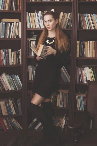 Krásná mladá dívka v černých šatech, stojící v knihovně s knihami — Stock fotografie