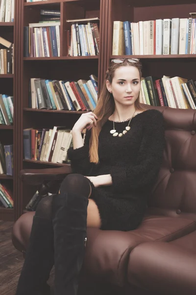Joven hermosa chica en un vestido negro sentado en un sofá de cuero en la biblioteca con libros —  Fotos de Stock