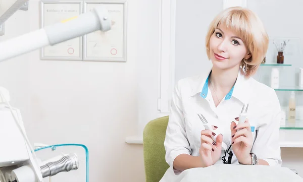  ofiste beyaz bir elbise içinde oturan ve makine aleti tutan genç sarışın bir kadın kalıcı güzellik uzmanı. Doktor. gülümseyen