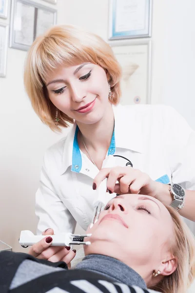 Mooie jonge blonde vrouw permanente schoonheidsspecialiste in een witte robe in het kantoor met de patiënt. Arts. glimlachen — Stockfoto