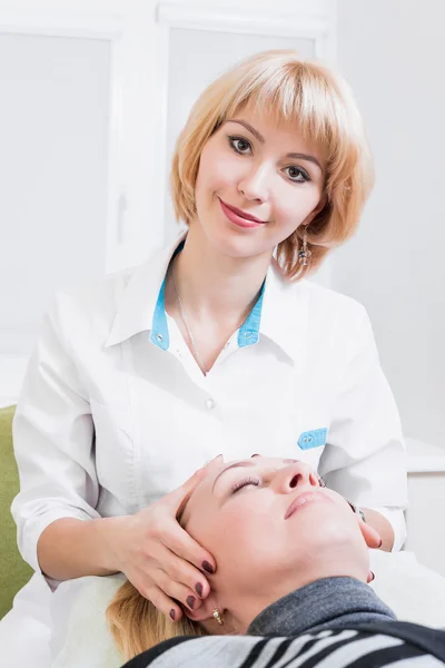 Femme blonde esthéticienne permanente en robe blanche au bureau avec massage facial patsientom.delaet. Docteur. souriant — Photo