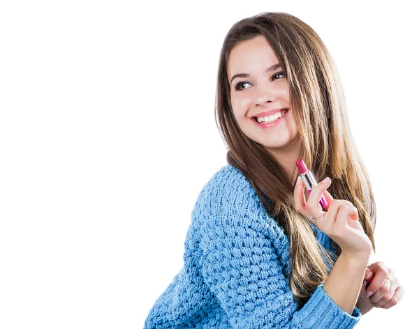 Beautiful young girl in a blue sweater standing on a white background and holding a red lipstick. Smiles. — Stock Photo, Image