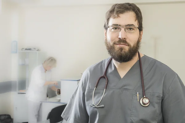 Médico barbudo vestindo óculos e um manto cinza fica em primeiro plano para o fundo para desfocar assistente — Fotografia de Stock