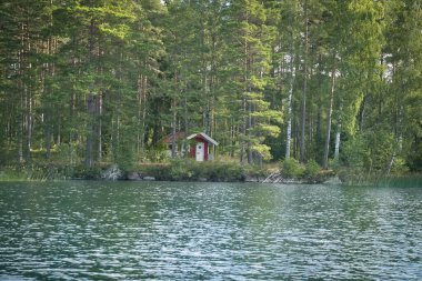 İsveç Gölü 'nde, sık ormanlarla çevrili kırmızı ahşap bir ev. Sudaki hafif yansımalar ve huzurlu bir atmosfer sakinliği ve doğayla bir bağ hissini iletir..