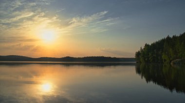 Altın güneş, orman ve bulutların çerçevelediği sakin göle yansıyor. Günün sonunda ışık, derinlik ve doğal ahenkle dolu sessiz bir an..