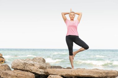 Yoga pozisyonunda kayalık bir sahilde duran bir kadın.