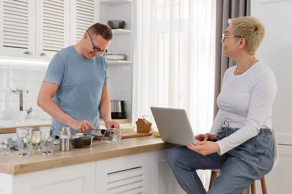 Fröhliches Paar mittleren Alters spricht beim Kochen und Arbeiten am Laptop über Küche — Stockfoto
