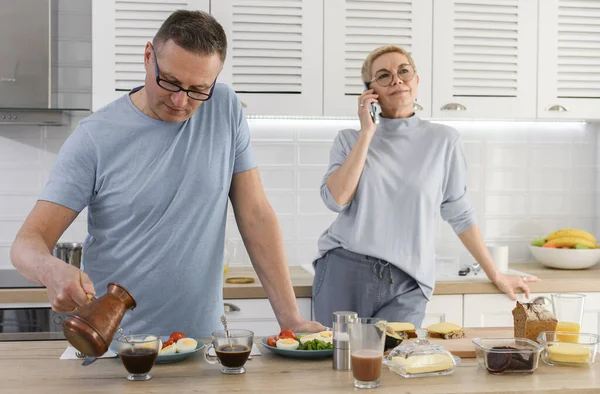 Senior Ehemann kocht Frühstück und Kaffee für seine Frau, während Frau telefoniert — Stockfoto