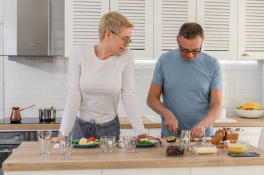 Orta yaşlı koca, mutlu yaşlı karısına mutfakta yemek pişirmesinde yardımcı olur, günlük kıyafetler giyer. Sevgi dolu olgun bir çift