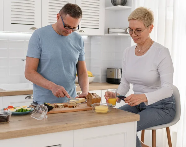 Gesundes, stilvolles Seniorenpaar, Frau schaut aufs Telefon, Mann steht neben ihr und kocht Frühstück, genießt die gemeinsame Zeit in der Küche — Stockfoto