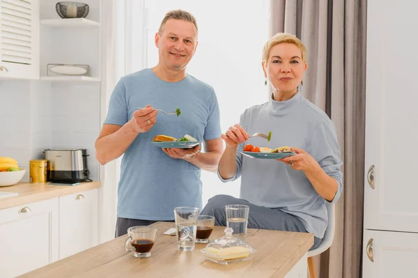 Gesunde reife Paare posieren vor der Kamera, während sie in der Küche gemeinsam gesundes Frühstück essen. Ehepaar mittleren Alters lebt zusammen — Stockfoto