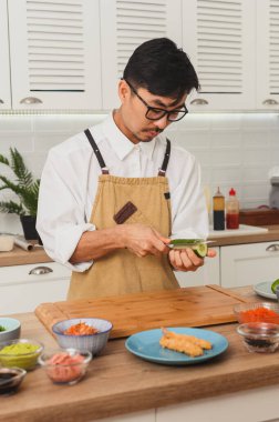 Asyalı şef suşi ruloları hazırlıyor, beyaz mutfakta salatalık doğruyor ve soyuyordu. Üniforma giyiyor.