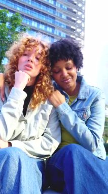 Açık havada oturan çok ırklı genç kadınlar, üzgün arkadaşını empati ve destekle rahatlatan, arkadaşlık ve şefkat ile dikey video formatında kentsel arka plan