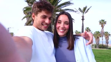 Palmiye ağaçlarının arka planında video görüşmesi yapan genç mutlu çift, gülümseyip kameraya el sallıyor, yaz boyunca neşeli iletişim ve teknoloji yaşam tarzı.
