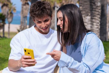 Genç bir adam ve kadın, deniz kenarındaki palmiye ağaçlarının altındaki ahşap bankta oturuyor. Sarı akıllı telefona birlikte bakıp, sosyal medyanın ve açık hava rahatlığının keyfini çıkarırken gülümsüyorlar. 