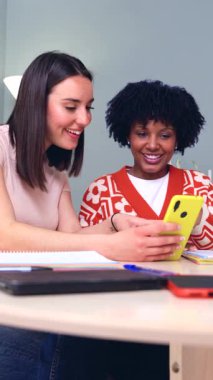 Kapalı alanda akıllı telefona bakarken gülümseyen iki farklı genç kadın, arkadaşlık, teknoloji, iletişim, öğrenme, bağlantı, mutluluk, çeşitlilik, takım çalışması ve eğitim kavramı.