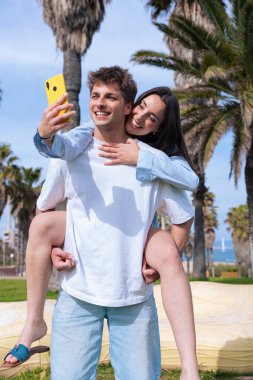 Palmiye ağaçlı genç mutlu çift, sarı akıllı telefon, yaşam tarzı, romantizm, eğlence, aşk, modern ilişki, çevrimiçi bağlantı, yaz eğlencesi.
