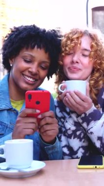 İki çok ırklı bayan arkadaş kahve dükkanında oturmuş gülüyor. Akıllı telefon, arkadaşlık konsepti, teknoloji, sosyal medya, çeşitlilik, bağlantı, yaşam tarzı, neşe ve modern gençlik..