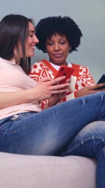 Neşeli Kafkasyalı ve Afro-Amerikalı kadınlar kanepeye oturmuş kırmızı bir akıllı telefona bakarken eğleniyorlar, kahkahaları paylaşıyorlar ve içeride kaliteli zaman geçiriyorlar. Dikey